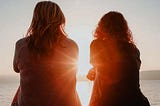 two female friends in silhouette looking at a sunset over water — Like in My Brilliant Friend