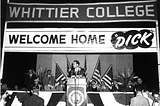 Black and white photo of Richard Nixon standing on stage in front of a podium with banners behind him reading “Whittier College” and “Welcome Home ‘Dick’.”