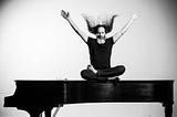 The author, sitting on top of a closed grand piano, arms outstretched, long hair flying.