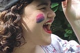 Lydia, a dark haired woman, wears a dark baseball cap, bright red lipstick, and the bisexual flag colors as face paint. She smiles big and proudly.