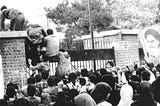 Black-and-white photo of male students climbing up Embassy gate as a crowd looks on