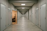A long institutional corridor with identical numbered doors, one standing open to reveal a warmly lit room with a desk and bookshelf. All other doors are closed.