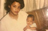 A sepia toned photo of a beautiful Black woman with light brown skin and voluminous black hair holding a baby.