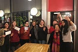 Photo of 8 women smiling, some holding up cards, some waving at camera. In a cafe, standing up behind table.