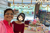 A woman and her daughter, around 15 years old, at a sweet shop. There are hampers of sweets behind them on a large table in the middle of the shop. There are festoons of marigold flowers decorating the shop. A Visicooler with Pepsi in it is at the far end. Another child is in the photo behind the mother and daughter, who is looking at the table with the hampers on it from the far end.