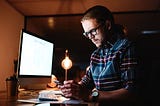 A man in front of a desktop. He keeps and looks at a mobile phone. He wears sunglasses.