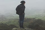 Photo of a man in a black hat and black coat and jeans standing on a ledge looking out over a foggy field