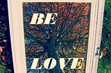 A mirror sitting on the Earth, framed in a cream frame, reflecting the trunk and colorful branches from the ground up inside the frame are the words “be love”