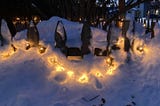 Fairy lights shining through snow outdoors hanging from a white picket fance