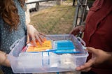 A woman hands a sample kit to a mom to collect dust from her home and cheek swabs for biological analysis.