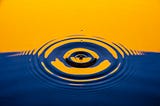 A close up of a water droplet piercing the stillness of a river reflecting orange and blue back hues.