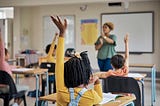 Classroom with a Black woman teacher at the front, and children raising their hands in the desks. Photo by pixdeluxe/Getty Images