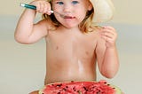 Little girl wearing hat eating a watermelon half with a spoon.