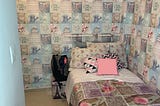 picture of a room that has wallpaper with squares of repeating quotes and patterns. quotes include “home sweet home” and “enjoy the little things” patterns include flowers, hearts, a watering can, a bicycle. also pictured is a bed. with similar repetitive square patterns with hearts and flowers, with differently patterned pillows on top. backpack sitting on top of a small ottoman that has a pattern with the Eiffel tower.