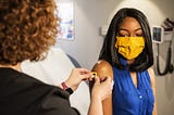 a Black woman wearing a blue sleeveless top receives a bandaid after receiving a vaccination from a white-appearing woman whose face is away from the camera