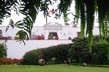 A white building, which is the front of Museo Larco, with flowers and bushes in front of it and the flag of Peru on top
