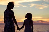 A young mother holds hands of her young daughter on a beach.