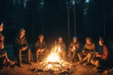 A group of campers huddled around a crackling campfire, their faces illuminated by its warm glow, surrounded by the dark shadows of the forest.