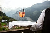 A food service worker offers a drink in a long-stemmed glass, overlooking a picturesque view of mountains.