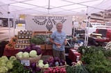 A woman farmer with gray hair at a booth full of vegetables at a Farm Stand.