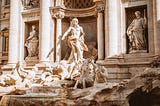 A view of the Trevi Fountain. Sculptures of roman gods and horses sculpted in white marble are touched by gentle ray of sun.