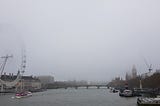 A view of a river with a bridge across it and half-shrouded Ferris wheel on one side and half-shrouded buildings in the distance due to fog