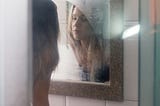 A young woman is leaned against a sink, contemplating her reflection in a foggy mirror.