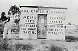 An old photograph of a man standing next to the side of a shed, on which is painted the words: “Bill Carter Mug Baker”