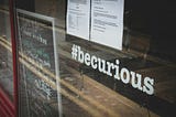 A picture of a window with an #becurious, two white papers that can’t read what is on them and a chalk board that can’t be read