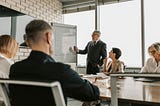 A man in a business suit at a whiteboard presenting something, while the rest of the team in business formal attire watches.