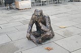 A bronze sculpture of a man sitting cross legged on the ground with his chin in one hand. It is nearly life sized.