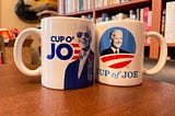 Two coffee mugs sitting on a dining table, one featuring a picture of Joe Biden inserted into the center of the Obama 2012 campaign logo with the text “Cup of Joe” written underneath, the other featuring a smiling portrait of Biden in his signature sunglasses, with the words “Cup o’ Joe” next to it.