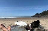 Author’s feet and surfboard and dog on the beach.