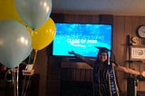 Author wears a graduation cap and blue-gold stole that reads “UCLA CLASS OF 2020”. She stands, arms open and palms up, in front of a TV screen mounted on the wall. The screen reads “Congratulations Class of 2020.”