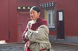 A tibetian woman standing against a red wall building. A sun is shining on her face and she is wearing a smile.