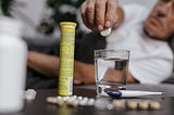 Someone is about to drop a tablet into a glass of water. There is a thermometer and medication on the table near them