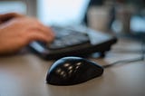 A computer mouse on a desk with hands on a keyboard slightly blurred in the background.