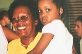 photograph of a black toddler looking warily at the camera, in the arms of an older black woman, who is smiling widely; the writer and her grandmother.
