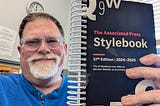 The author, John Carney, holding a spiral-bound 2024–26 Associated Press Stylebook.