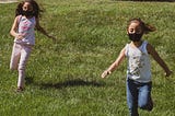 two girls wearing cloth face masks and playing on a green grass field during daytime