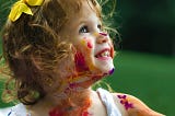 A two or three year old girl smiling at her parent or caregiver with paints on her face and dress.