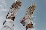 A person’s cuffed jeans and white converse with red heart designs on them raised toward a light blue sky with wispy clouds