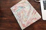 a rainbow paint pour soft cover notebook diary next to a macbook keyboard plugged into the charger