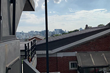 In the foreground is a residential brick house. To the left are concrete stairs. High-rise buildings stand in the distance against the backdrop of a blue sky, with some clouds within it.