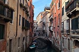 Photo of a narrow Venice canal and bridge with red, white and yellow houses on either side and a boat moored in the foreground.