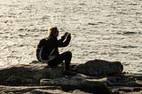 Photograph of a man with a backpack, mostly silhouetted, sitting on a rock with the sea in the background, taking a photograph on his phone.
