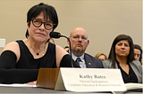 Kathy Bates with short dark hair, wearing glasses, a black top and black compression sleeves, sitting at a table with a microphone testifying before Congress
