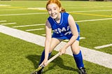 A little red-headed girl, age 11, in a blue field hockey uniform, posing for a picture on the field.