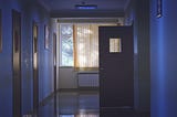 Dark hospital hallway with one door open with a window at the end covered by curtains