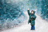 A child standing on a dirt road catches a colorful leaf falling with the snow.